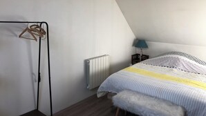 A bedroom with a bed, a radiator, a wooden hanger rack, a fluffy white bench, and a nightstand with a lamp.
