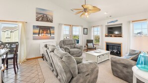 A spacious living room with a gray sectional sofa, a recliner, and a coffee table. There is a fireplace, a TV mounted on the wall, and large windows offering a view of the outside.