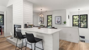 A modern kitchen with a white island, black bar stools, a fireplace, and a dining area. - Inspect2Sea: Pool, Hot Tub | Walk to Downtown Duck (Duck)