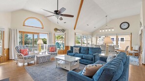 A spacious living room with a blue sofa, white coffee table, and wicker chairs. There is a ceiling fan, large windows, and a clock on the wall. - Lighthouse Jewel: Private Pool/Hot Tub/Sound Views (Corolla)