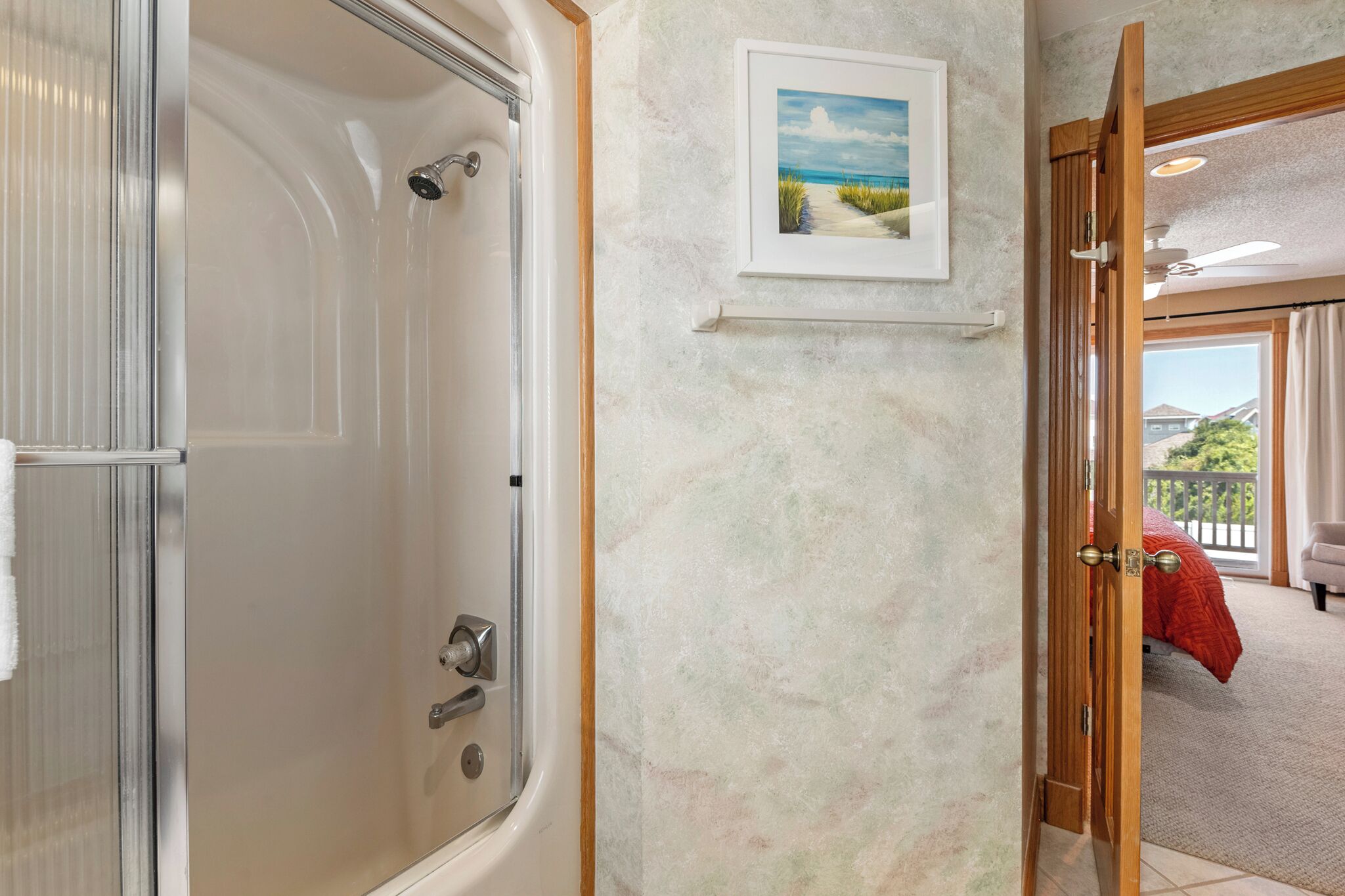 Une salle de bain dotée d’une douche à parois de verre, d’un tableau encadr é accroché au mur et d’une porte ouverte donnant sur un balcon où est posée une couverture rouge.
