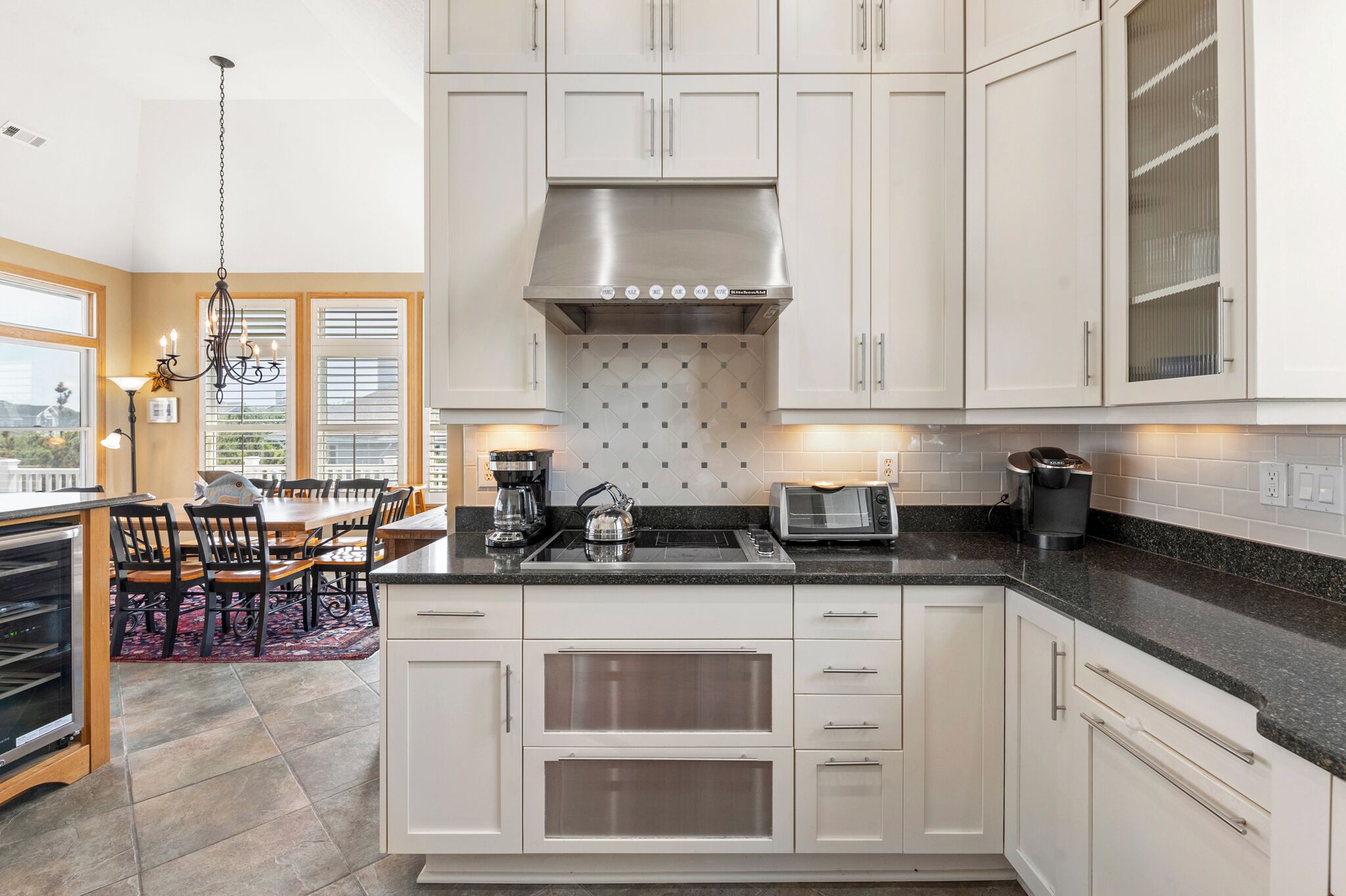 A modern kitchen with white cabinets, a stainless steel range hood, a coffee maker, a toaster, and a dining area with a table and chairs.