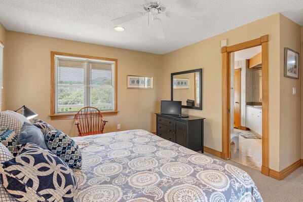 A bedroom with a bed, a desk, a chair, a ceiling fan, a window with blinds, and a door leading to a bathroom.