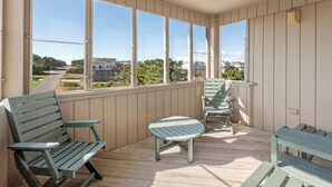 A covered porch with wooden benches and a small table, large windows offering a view of a residential street. - Chirality: Oceanfront | Direct Beach Access (Duck)