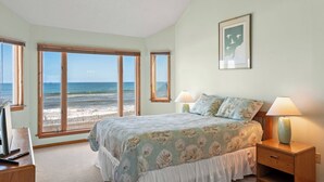 A bedroom with a large window overlooking a beach, a bed with floral bedding, two bedside lamps, and a small wooden nightstand. - Chirality: Oceanfront | Direct Beach Access (Duck)