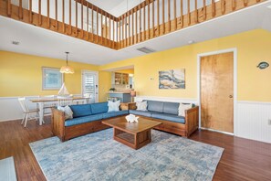 A living room with a blue sectional sofa, wooden coffee table, and a dining area with a white table and chairs. - It's A Shore Thing: Ocean Views | Hot Tub | Dogs OK (Rodanthe)
