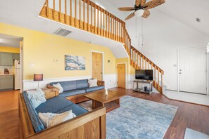 A spacious living room with a wooden staircase, a sofa set, a flat-screen TV, and a ceiling fan.