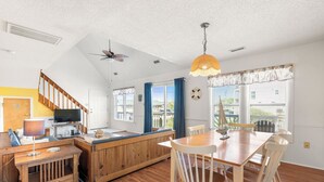 A spacious kitchen with a wooden island, a dining area with a table and chairs, a ceiling fan, and large windows with curtains. - It's A Shore Thing: Ocean Views | Hot Tub | Dogs OK (Rodanthe)