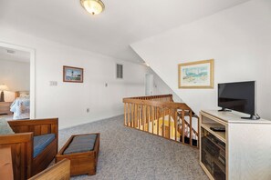 A spacious living room with a high ceiling, a wooden staircase, a television, and two framed artworks.
