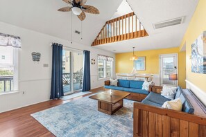 A modern living room with a wooden ceiling, a blue sofa, a coffee table, and a dining area with a yellow wall. - It's A Shore Thing: Ocean Views | Hot Tub | Dogs OK (Rodanthe)