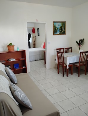 Een woonkamer met een bank, een eettafel met stoelen, een klein boekenrek en een deur die naar een slaapkamer leidt.