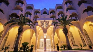 Ein Hotel mit Eingangsbereich, einem zentralen Bogen, Palmen und einer symmetrischen Fassade mit Balkonen.
