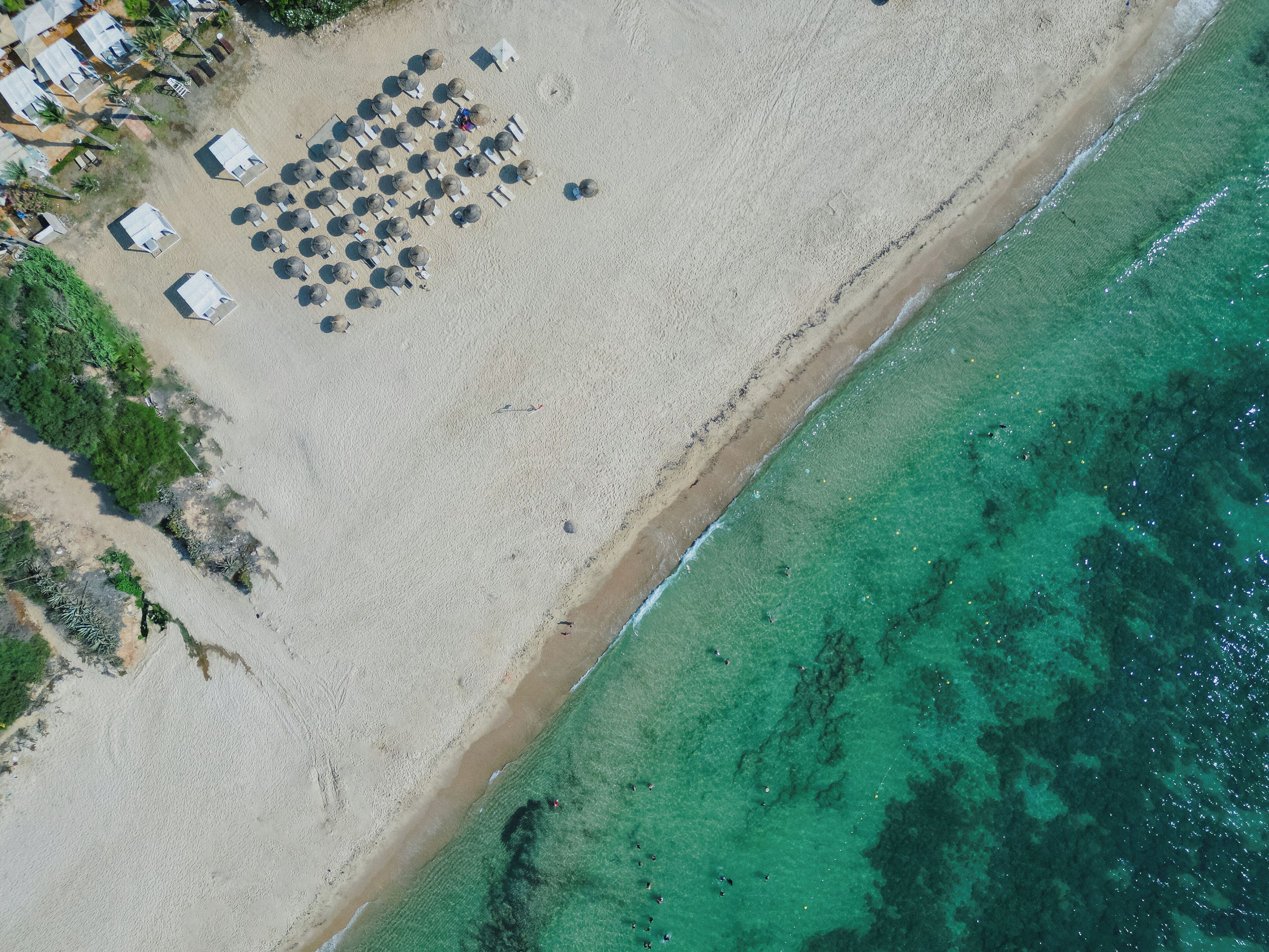An aerial view of a beach with rows of beach huts and umbrellas, a clear shoreline, and a calm sea.