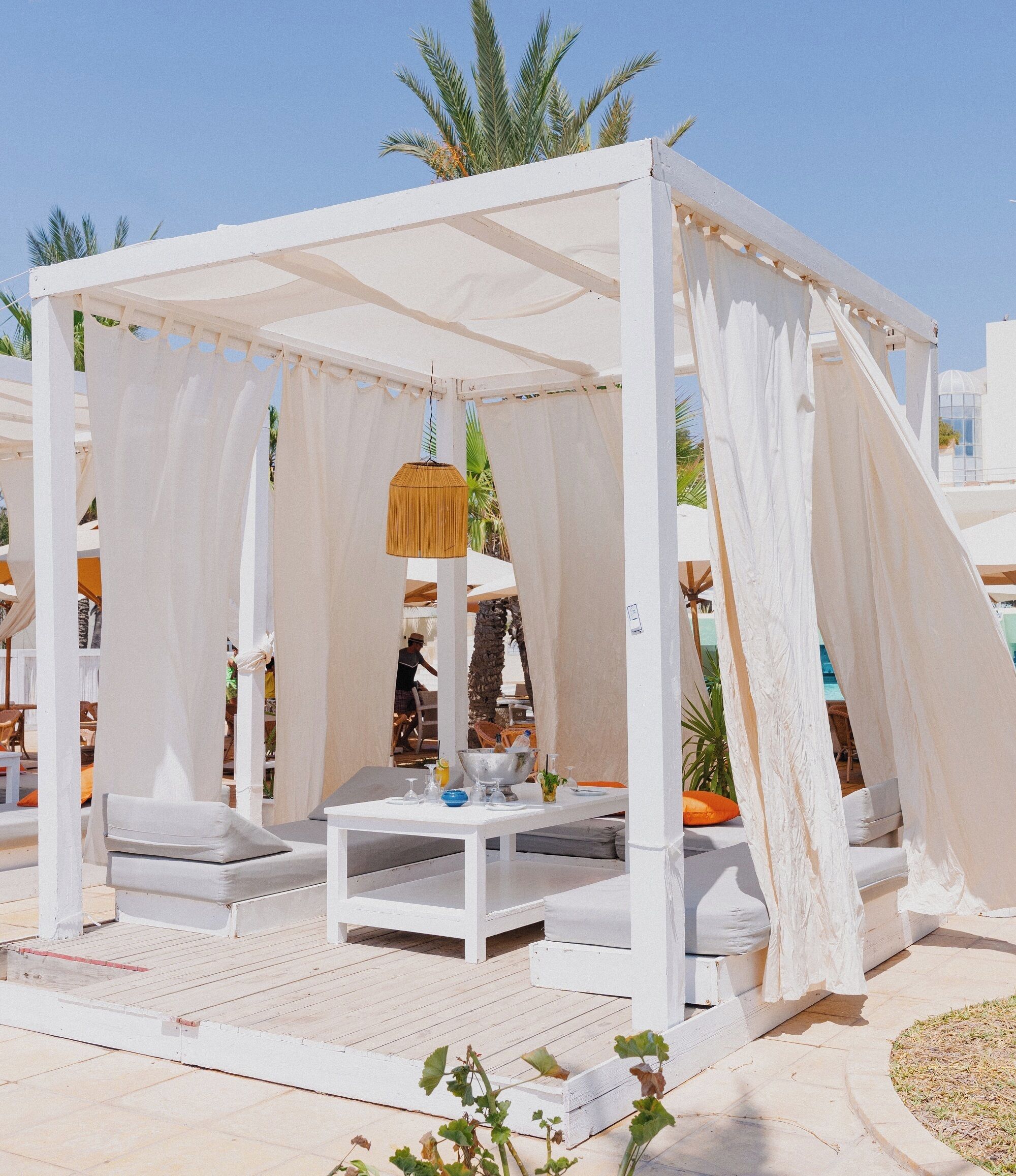 Un espacio al aire libre con asientos blancos, una mesa y cojines, rodeado de cortinas blancas y palmeras.