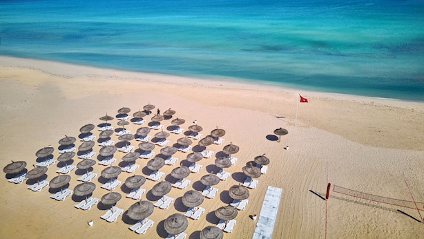 Ein Strand mit Reihen von Liegestühlen und Reetdachschirmen, einem Volleyballnetz und einer Flagge.