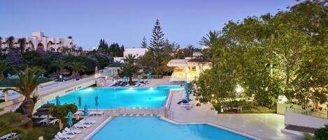 A large outdoor swimming pool with lounge chairs and umbrellas, surrounded by greenery and buildings.