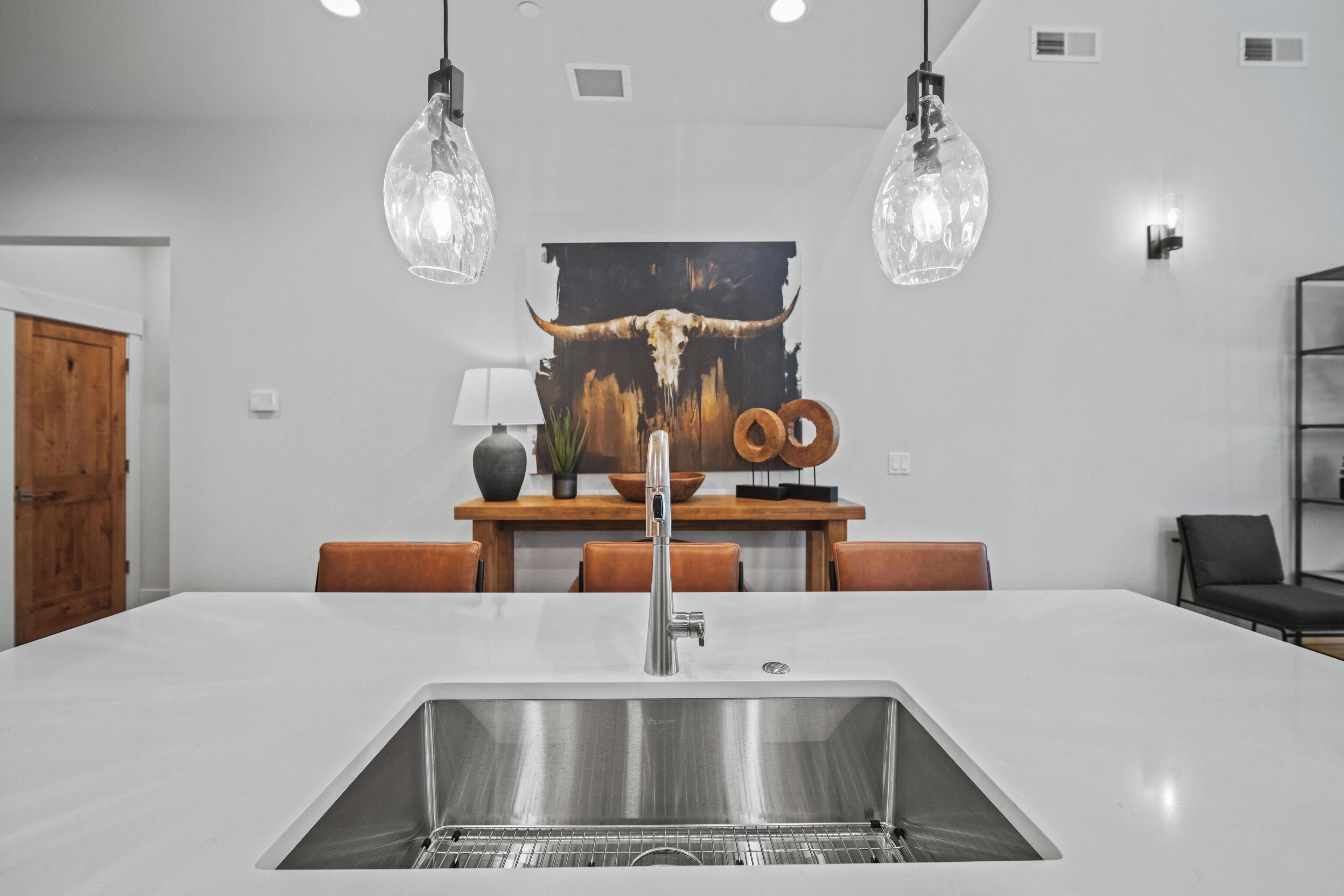 A modern kitchen with a large island, stainless steel sink, pendant lights, and a rustic wall art piece.