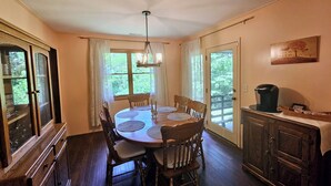 A dining area with a wooden table and chairs, a chandelier, and a view of greenery through the windows. - The Marigold Cottage: Serene Mountain Getaway w/ Fire Pit, Large Yard (Highlands)