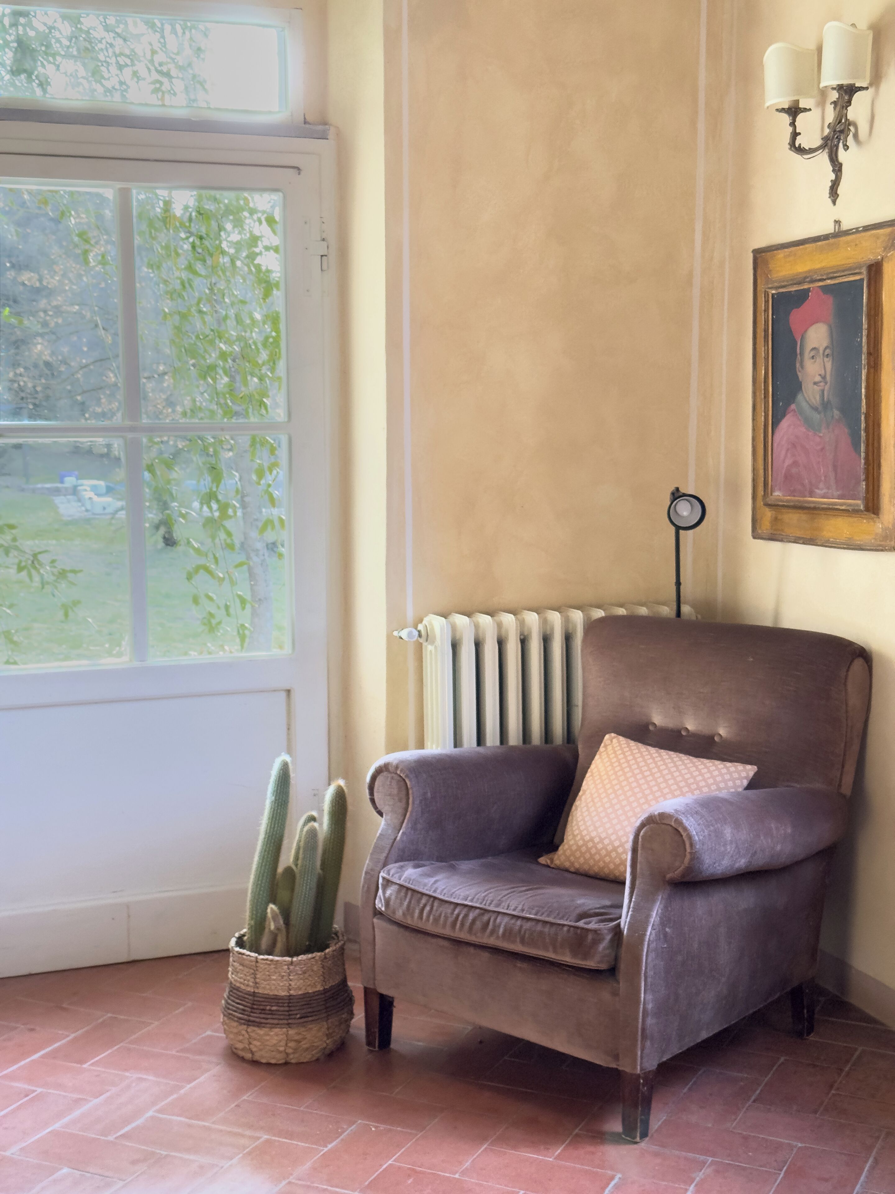 Una habitación con un radiador, una silla con un cojín, un cactus en una cesta y un retrato enmarcado en la pared.