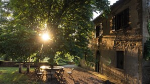 Un espacio de estar al aire libre de estilo rústico con una mesa y sillas de piedra, rodeado de abundante vegetación y un edificio histórico con ventanas de arco.