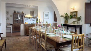 Un comedor con una mesa de madera dispuesta para cuatro personas, rodeada de sillas de madera. Se ve una cocina al fondo con un armario de madera y diversos utensilios.