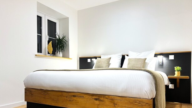 Ein modernes Schlafzimmer mit einem Holzbettgestell, weißer Bettwäsche, einem kleinen Nachttisch mit Pflanze und einem Fenster mit Blick.