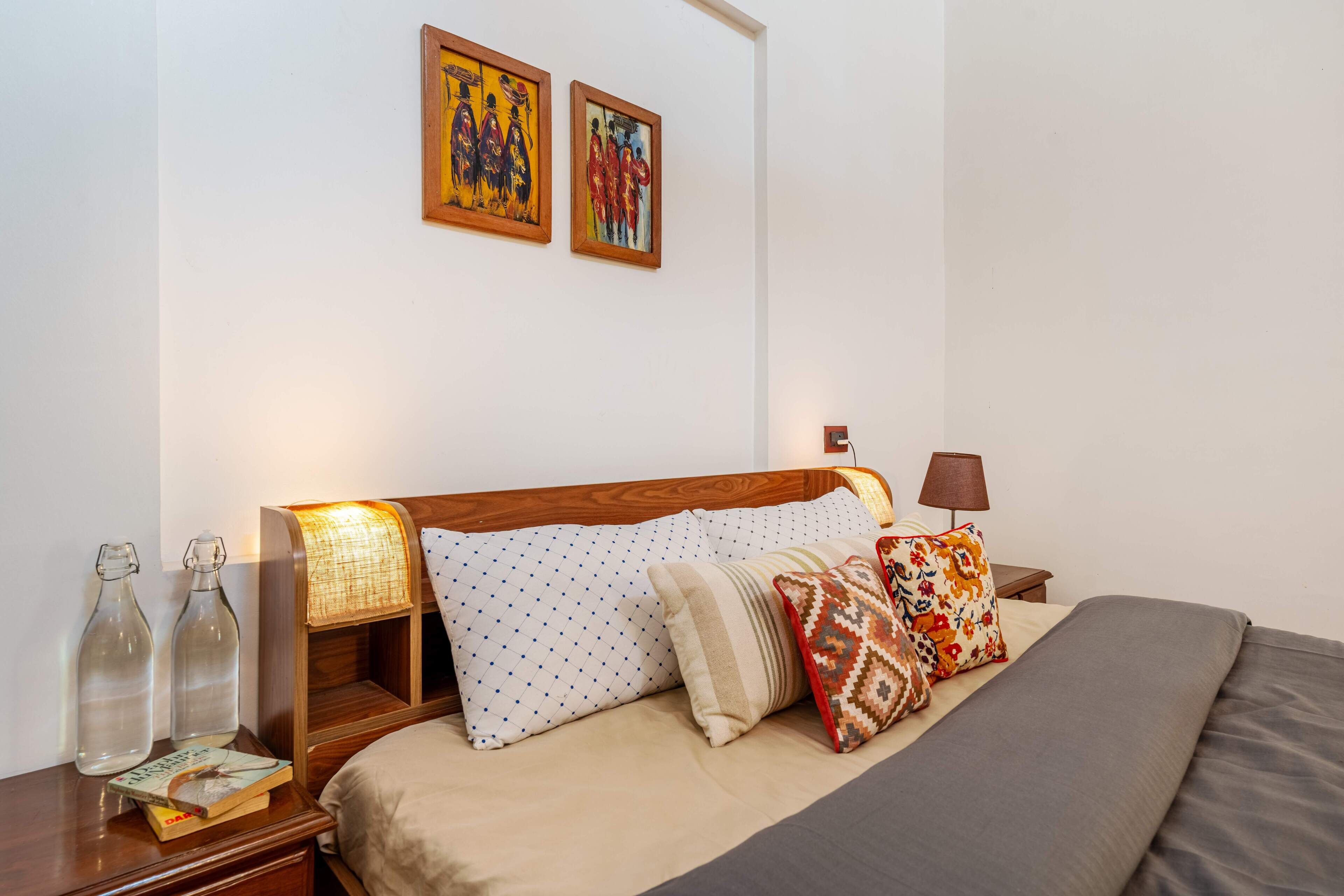 Una cama bien hecha con cabecera de madera, dos lámparas de noche y una mesita de noche con libros y botellas.