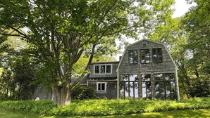 Ein zweigeschossiges Haus mit Giebeldach und großen Fenstern, umgeben von üppigem Grün und einem gepflegten Rasen.