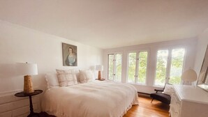 Ein Schlafzimmer mit einem großen Bett, zwei Nachttischen, einer Kommode und einem Fenster mit Blick auf Bäume.