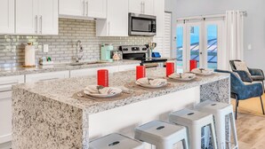 A modern kitchen with white cabinets, a granite countertop, a microwave, and a stove. There are four red candles on the counter, and a blue chair is visible in the background. - Seahorse Suites #4. A Perfect Beach Getaway! (Sanibel)