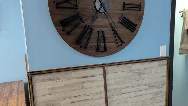 Une grande horloge murale en bois, dotée de chiffres romains et d’un balancier, fixée à un mur bleu.
