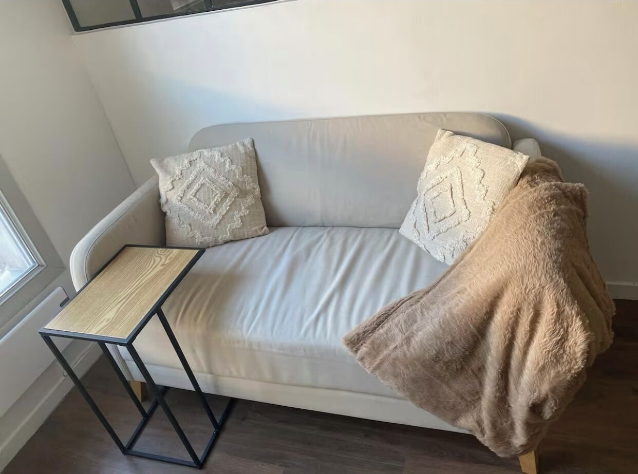 A sofa with two patterned pillows, a wooden side table, and a beige throw blanket.