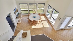 A spacious room with a round dining table surrounded by chairs, large windows offering a view of the outdoors, and a staircase leading to another level.