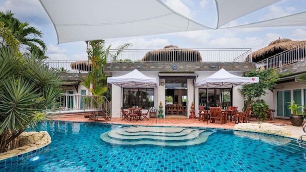 A swimming pool with tiled steps, surrounded by outdoor seating and umbrellas, and a building with a thatched roof and a balcony. - Sahaswara Luxury Villa Pattaya (Bang Lamung)