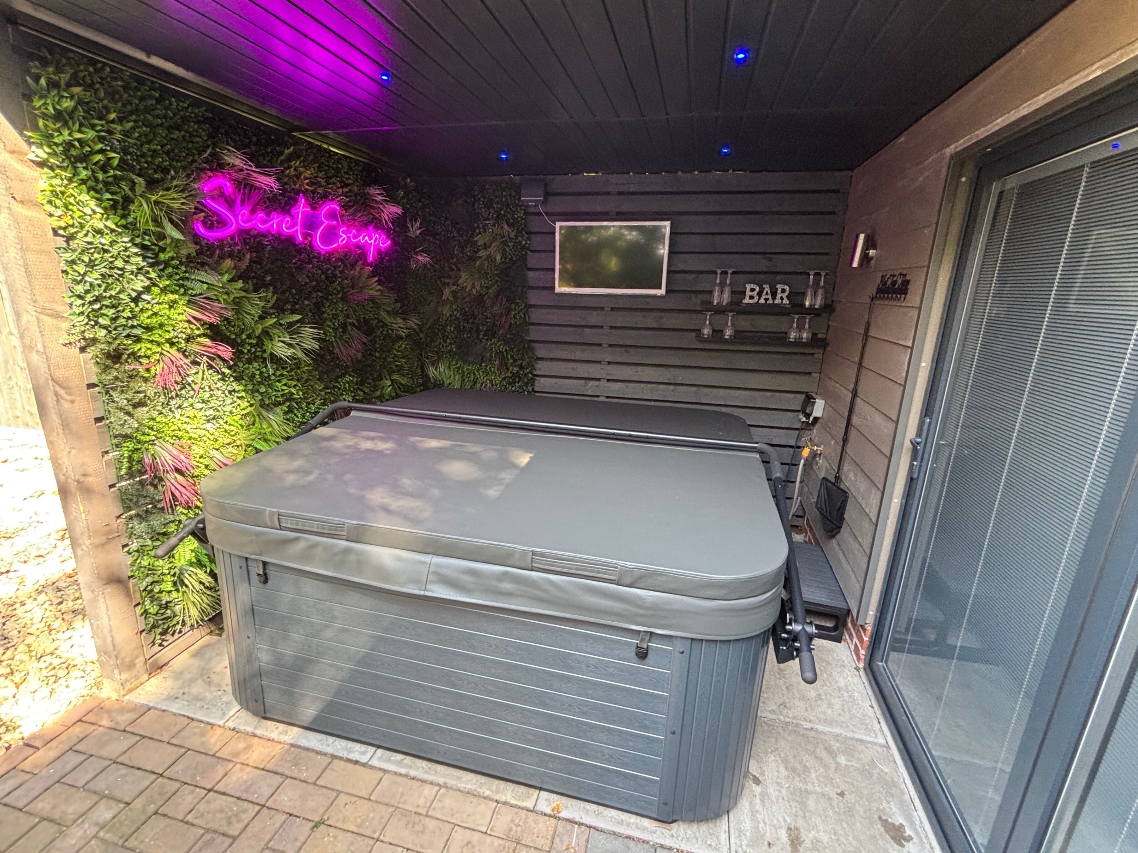 Een hottub met een houten terras, omgeven door weelderige begroeiing en een neonreclame.