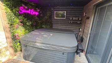 Een hottub met een houten terras, omgeven door weelderige begroeiing en een neonreclame.