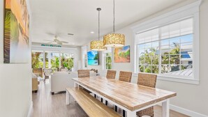 A spacious dining area with a large wooden table, wicker chairs, and a bench. There are two pendant lights above the table. The room has large windows with a view of palm trees and a building. - Anna Maria Home with Pool and Rooftop Deck! 5-min Walk to Beach - Coconut Breeze (Anna Maria)