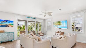 A spacious living room with a large window, a ceiling fan, and a view of the outdoors. - Anna Maria Home with Pool and Rooftop Deck! 5-min Walk to Beach - Coconut Breeze (Anna Maria)