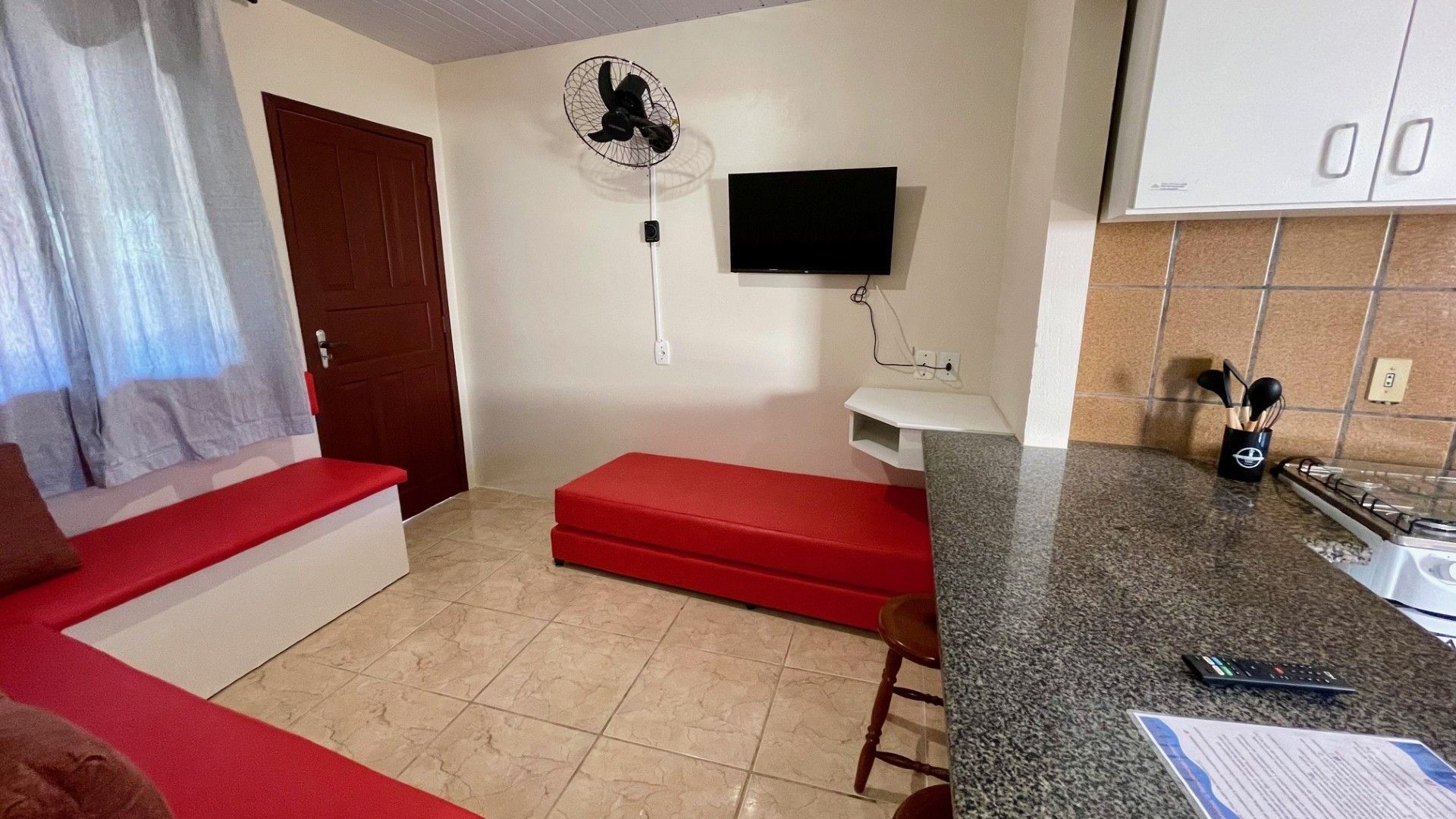A compact living space with a red sofa, a small red bench, a TV mounted on the wall, a kitchen area with a countertop and stove, and a ceiling fan.
