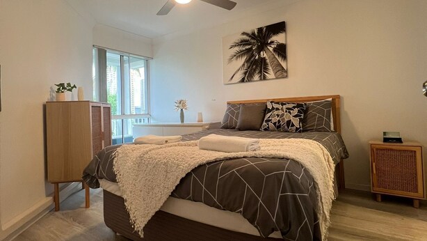 Un dormitorio con cama, ventilador de techo, ventana y un cuadro en la pared.