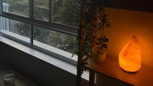 Una habitación con un ventanal grande, una lámpara de sal, una planta en maceta y un cartel en la pared.