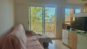 Una habitación con sofá, televisor y balcón con vistas a palmeras y al mar.
