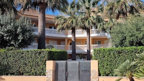 Un hotel de varios pisos con balcones, palmeras y un seto bien cuidado al frente.