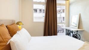 Une chambre d’hôtel comprenant un lit, un bureau, une chaise, une lampe, une fenêtre avec des rideaux et un balcon avec vue.