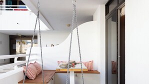 Un balcon avec un fauteuil suspendu, une table en bois avec un coussin et une porte-fenêtre coulissante.