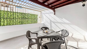 Een zonnig terras met een tafel en stoelen, uitzicht op groen achter een metalen hek, en een witte muur.