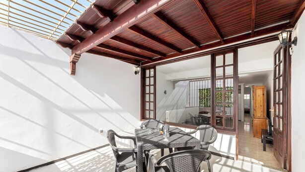 Een zonnig terras met een tafel en stoelen, een houten pergola en uitzicht op een kamer met een bed en gordijnen.