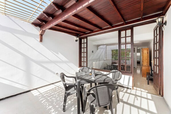 Un patio soleado con una mesa y sillas, una pérgola de madera y vistas a una habitación con cama y cortinas.
