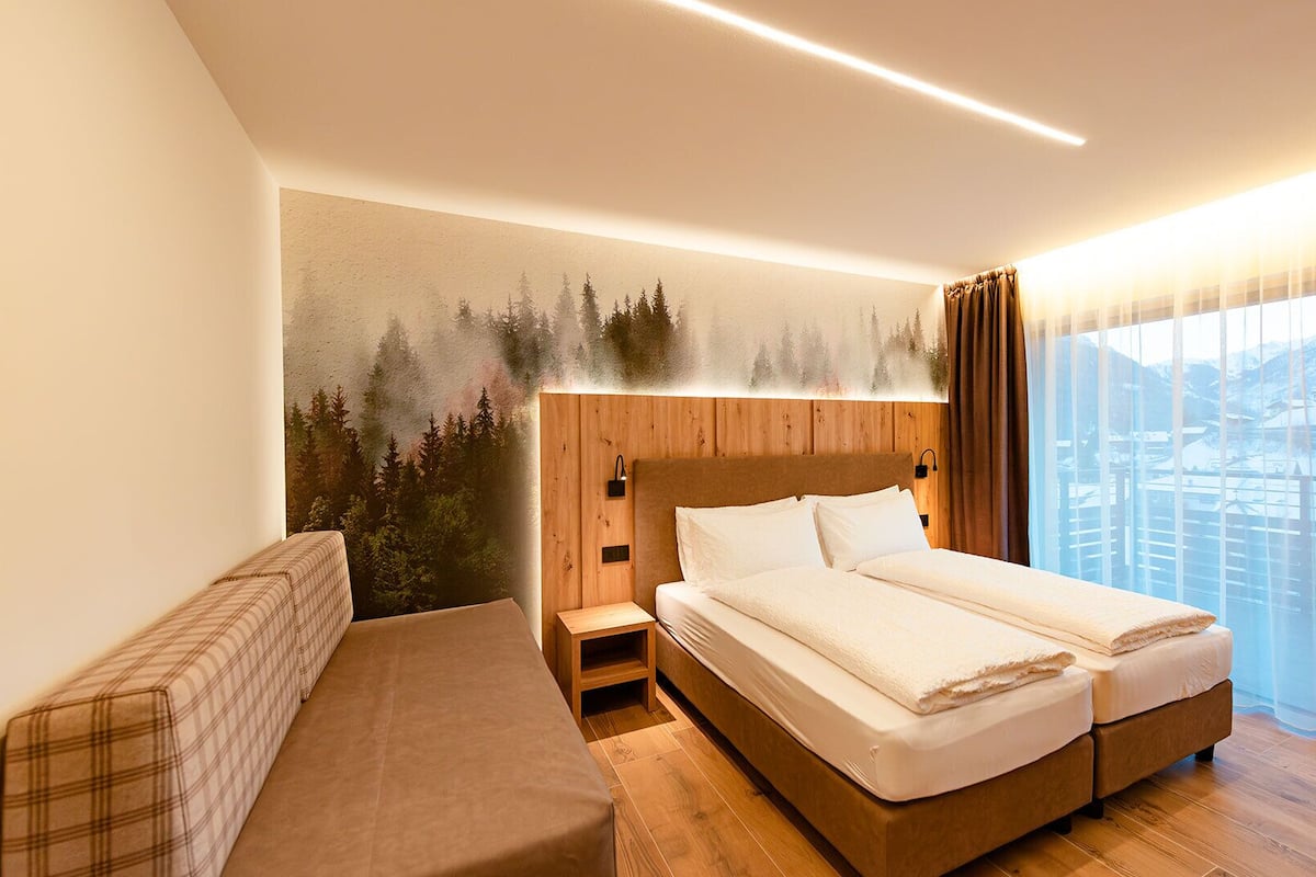 A hotel room with a bed, a bench, and a view of a snowy landscape through large windows.