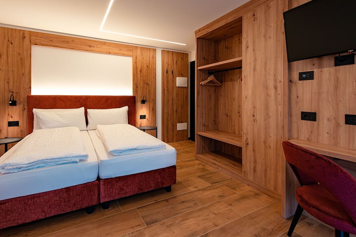 A double bed with white linens, a red upholstered headboard, and a wooden wardrobe with a mirror.